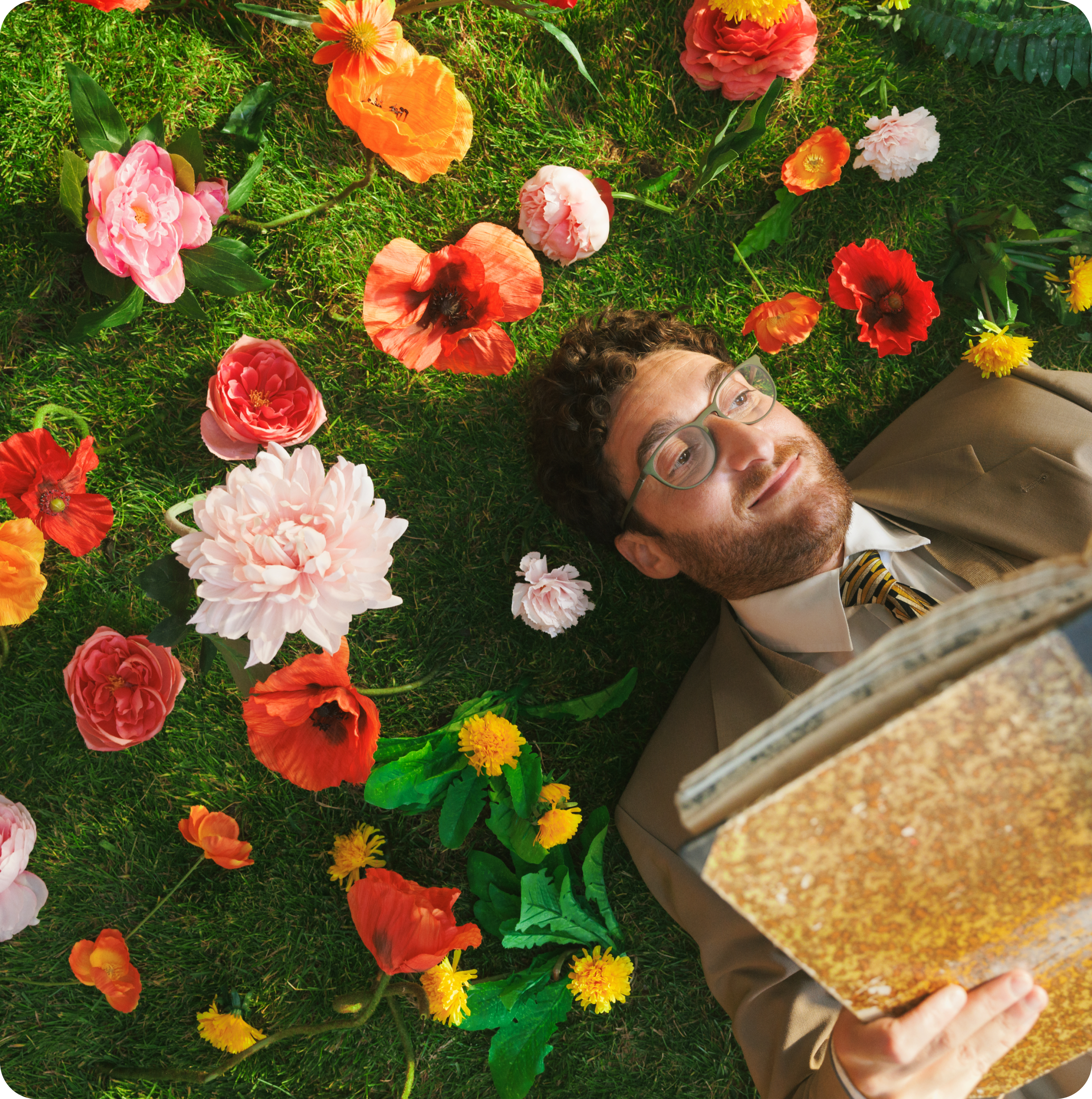 A man with curly hair and glasses, dressed in a beige suit and striped tie, is lying on green grass surrounded by vibrant flowers.